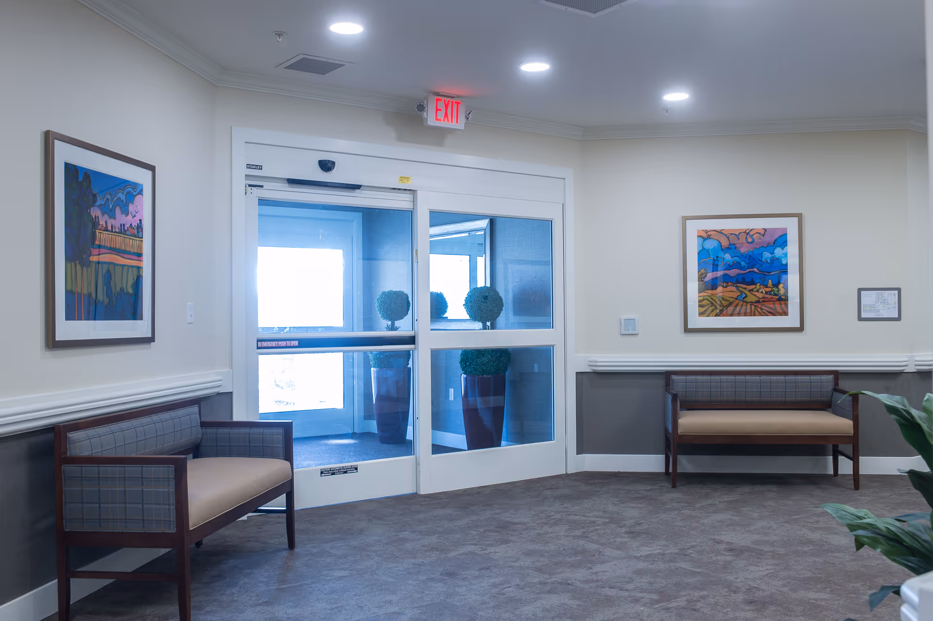 Interior view of a waiting area near an exit door in a senior living facility. The space features two wooden benches with cushioned seats and backs, framed colorful landscape paintings on the walls, and a glass automatic door with an illuminated red exit sign above it. There are also two large potted plants visible through the glass door.