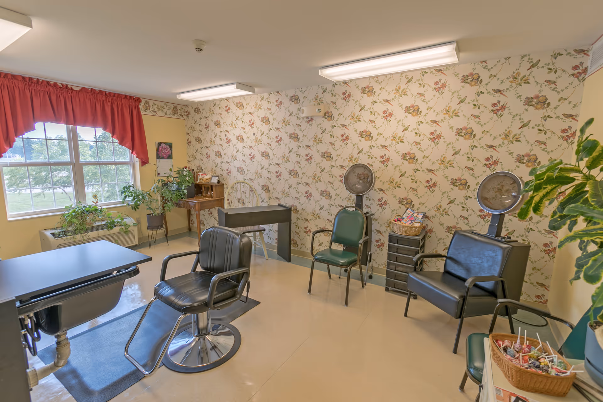 A cozy hair salon room with floral wallpaper and a large window with red curtains. The room contains a black salon chair in front of a sink, two green chairs with hair dryers attached, a small wooden desk with a chair, and several potted plants. There is also a basket of lollipops on a small table.