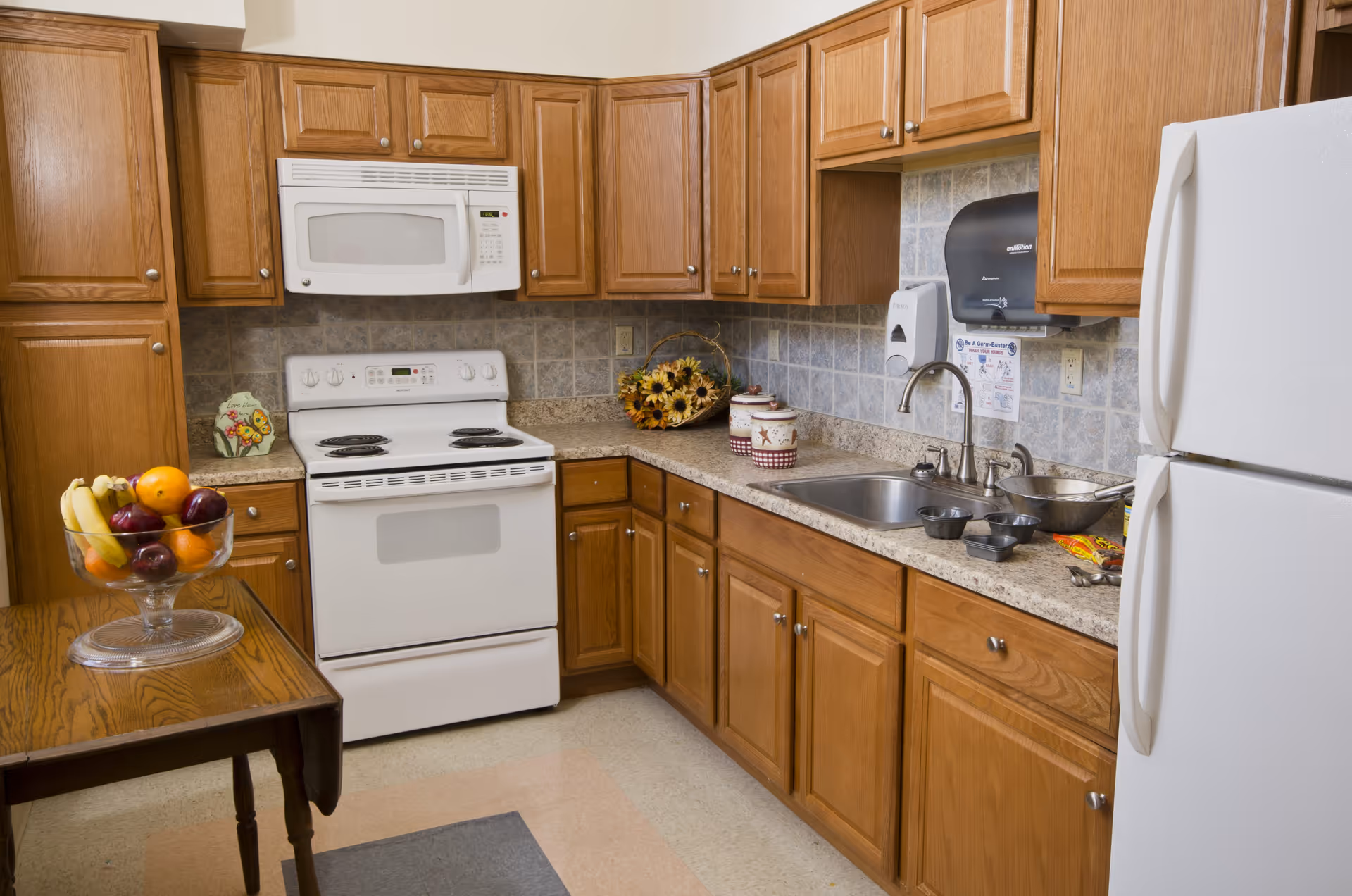 A kitchen with wooden cabinets, a white electric stove with an oven, a white microwave above the stove, a double sink with a faucet, and a white refrigerator. There is a small wooden table with a glass bowl of assorted fruit including bananas, oranges, and apples. The backsplash is tiled in a light gray pattern, and there are various kitchen items on the countertop including a basket of sunflowers and some containers.
