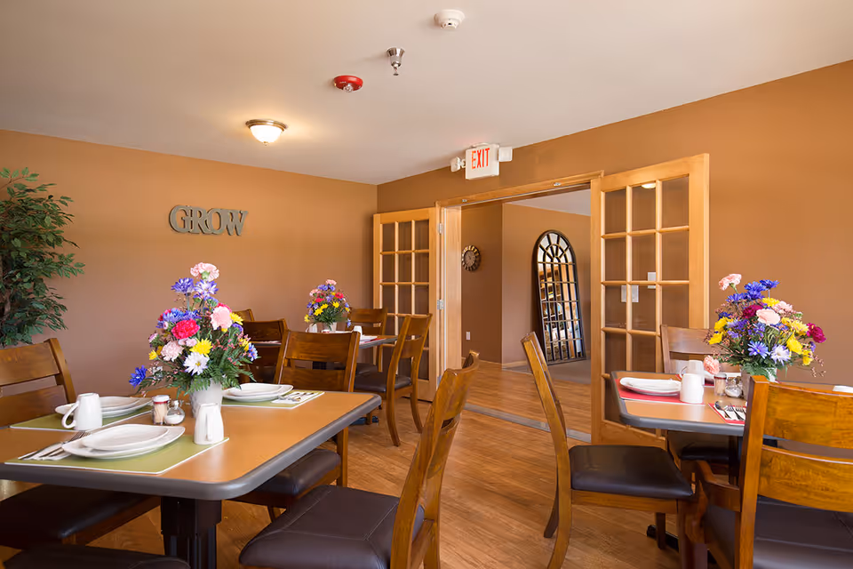 A dining room with wooden tables and chairs, each table set with plates, cups, and colorful flower arrangements. The walls are painted brown, and there is a decorative word 'GROW' mounted on one wall. Double wooden doors with glass panes lead to another room with a large arched mirror and a wall clock.