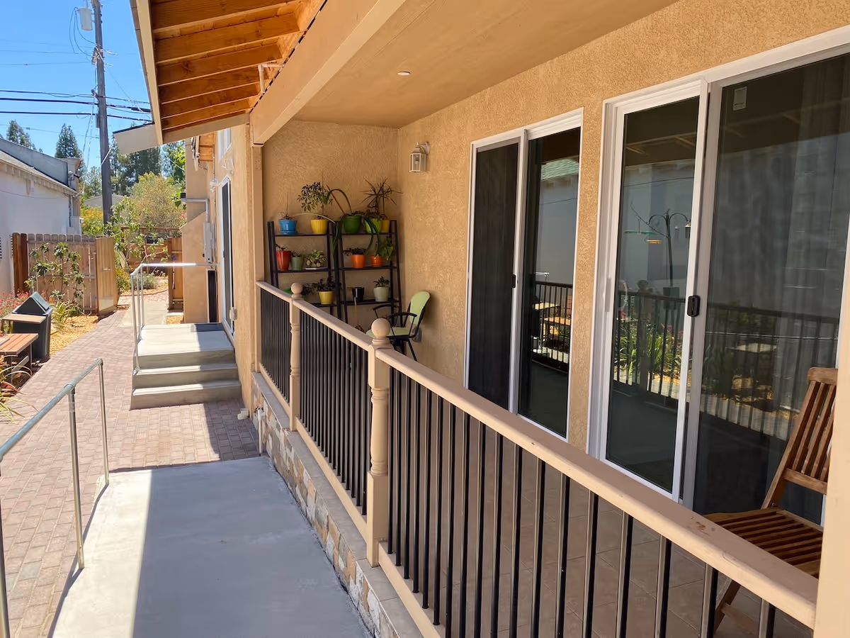 Covered exterior walkway beside a senior living building with sliding glass doors, a railing, potted plants on shelves, and steps and a ramp leading to a garden.