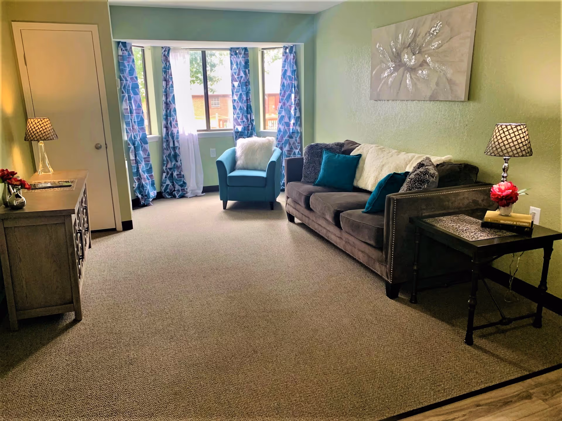 Sunlit living room with a brown couch, teal armchair by a window with patterned curtains, side tables with lamps, and green walls.