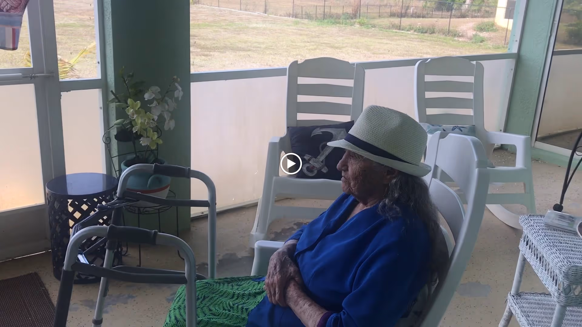 An elderly woman wearing a white hat and blue clothing sits on a white plastic chair on a covered patio. There are additional white plastic chairs, a walker, a small table with a plant, and a wicker shelf nearby. The patio overlooks a grassy outdoor area with a fence in the background.