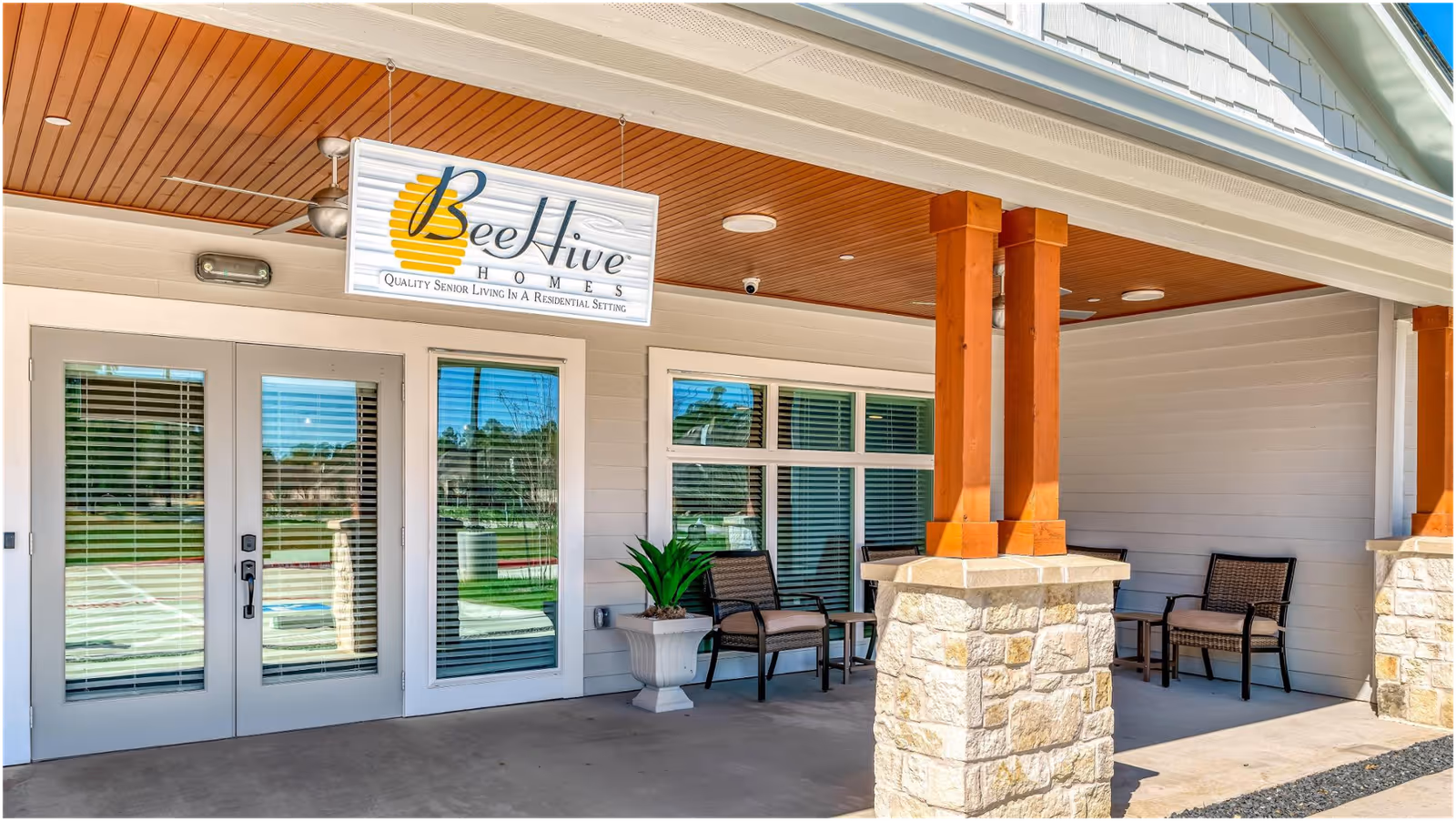 Entrance area of BeeHive Homes of Spring featuring a covered porch with stone and wooden pillars, two sets of glass doors with blinds, a potted plant, and several chairs arranged for seating.