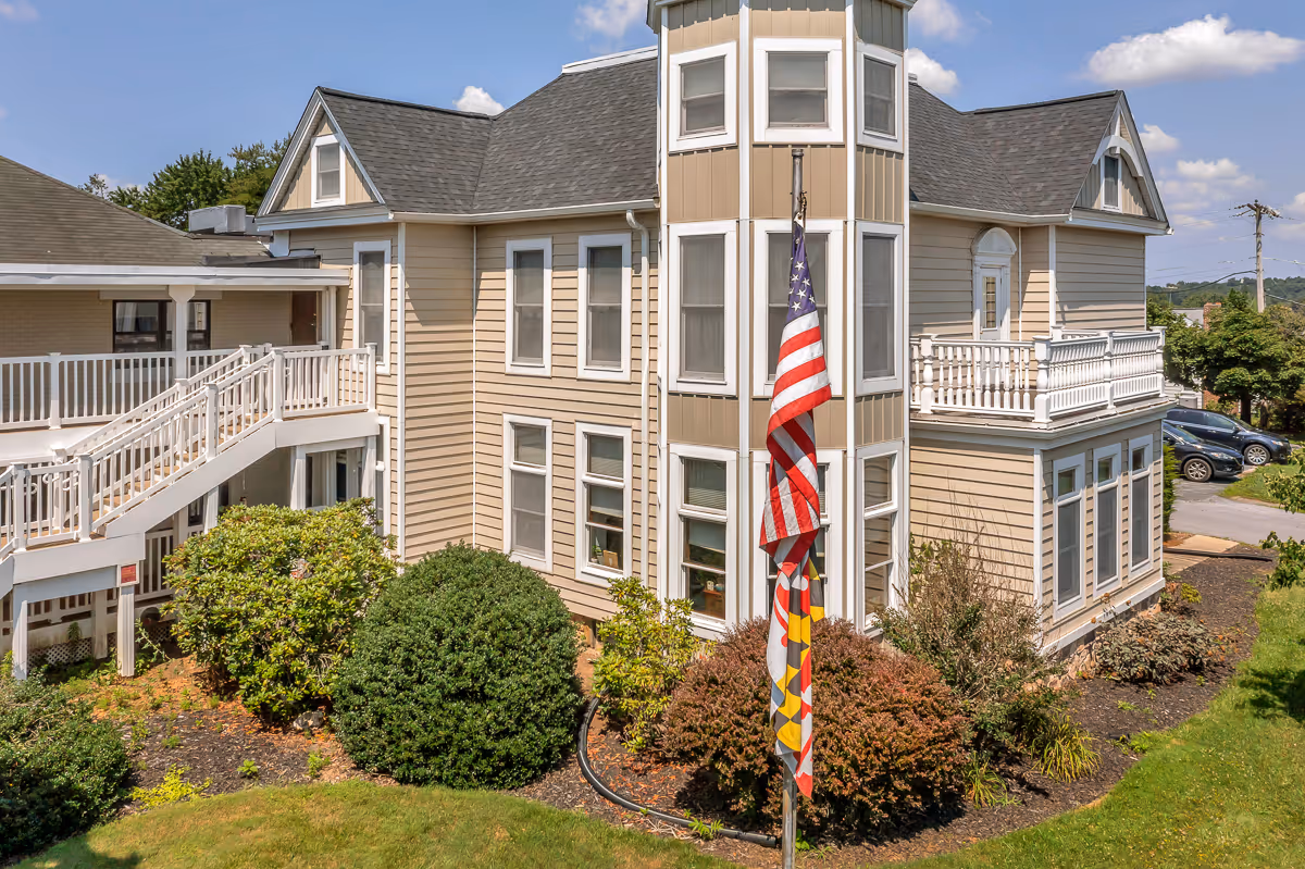 Exterior view of a beige multi-story senior living building with white trim, a flagpole displaying the U.S. and Maryland flags, and landscaped shrubs.