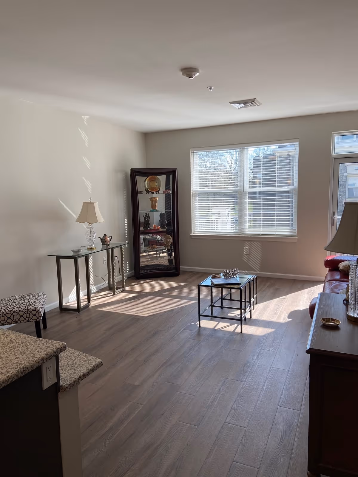 A bright living room with large windows letting in natural light. The room features a glass display cabinet with decorative items, a glass console table with a lamp and small decor, a small coffee table with a decorative piece, a red leather sofa partially visible, and wood-look flooring.