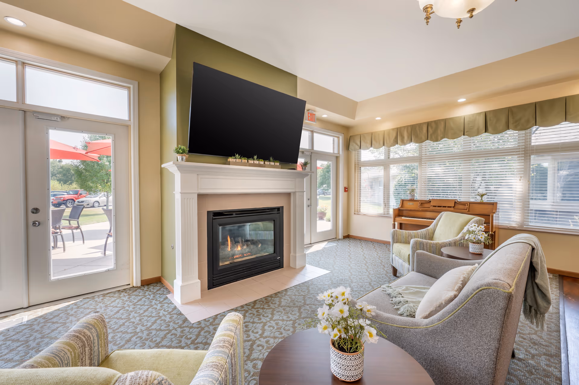 A bright and cozy living room area in a senior living facility featuring a fireplace with a large flat-screen TV mounted above it. The room has comfortable seating including armchairs and a sofa, a small round table with a flower pot, large windows with blinds and valances, and a piano in the corner. There are two glass doors leading outside to a patio area with chairs and umbrellas.