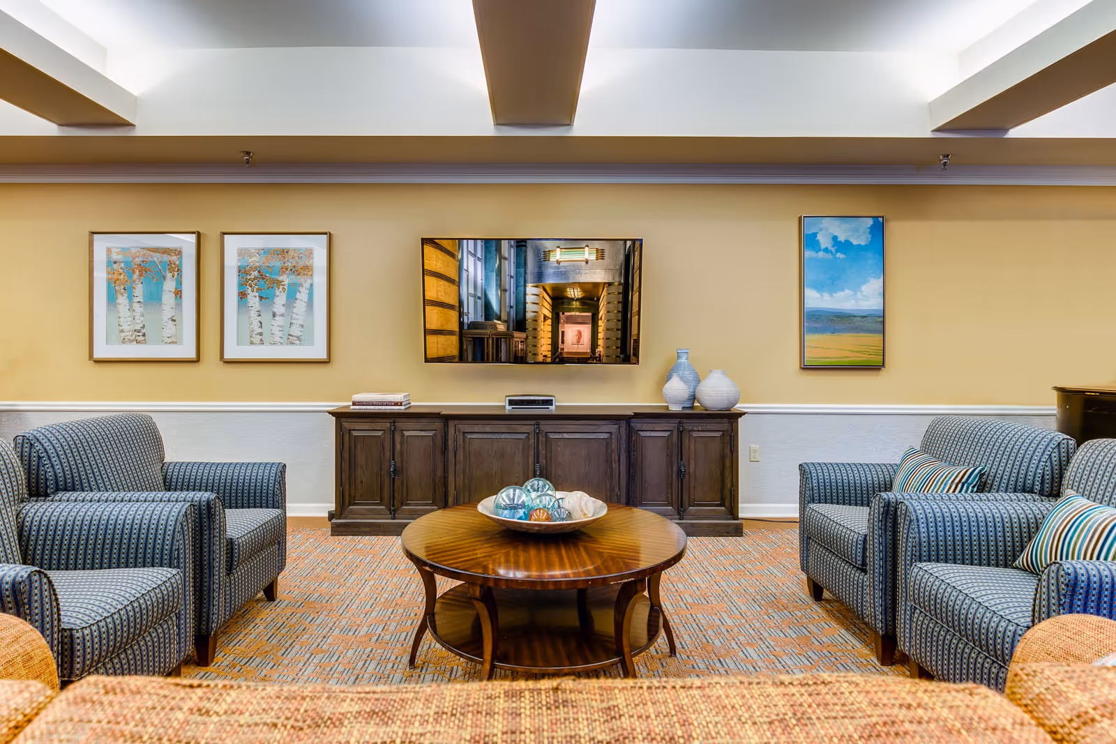 Comfortable common sitting area with upholstered armchairs, a round coffee table, a wall-mounted TV and framed artwork.