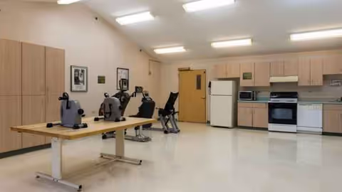Bright communal rehab room with a long table, exercise machines, and a kitchen in the background.