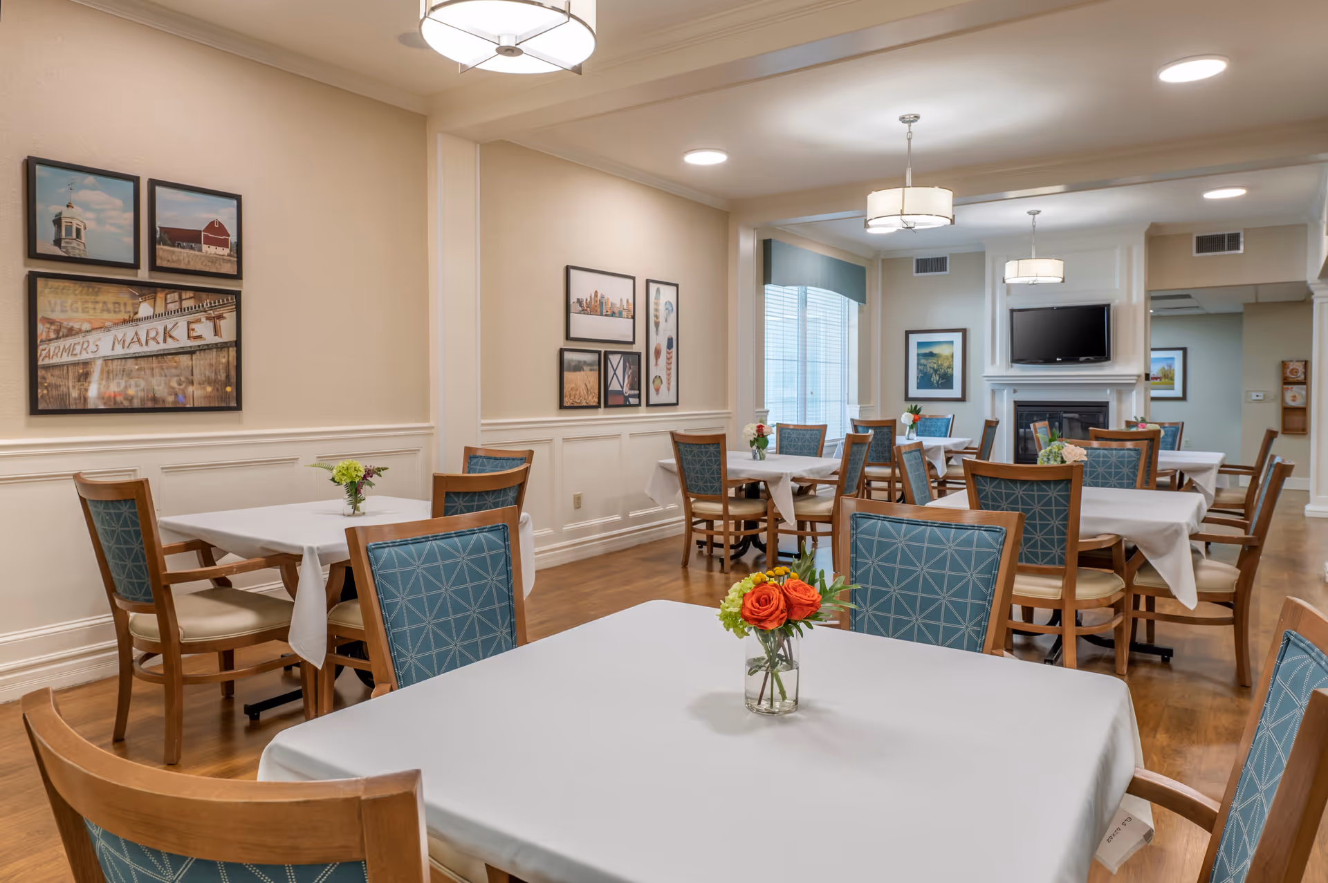 A dining room in a senior living facility with multiple tables covered in white tablecloths, each adorned with small flower arrangements. The room features wooden chairs with blue patterned cushions, framed pictures on the beige walls, a fireplace with a mounted TV above it, and ceiling lights providing warm illumination.