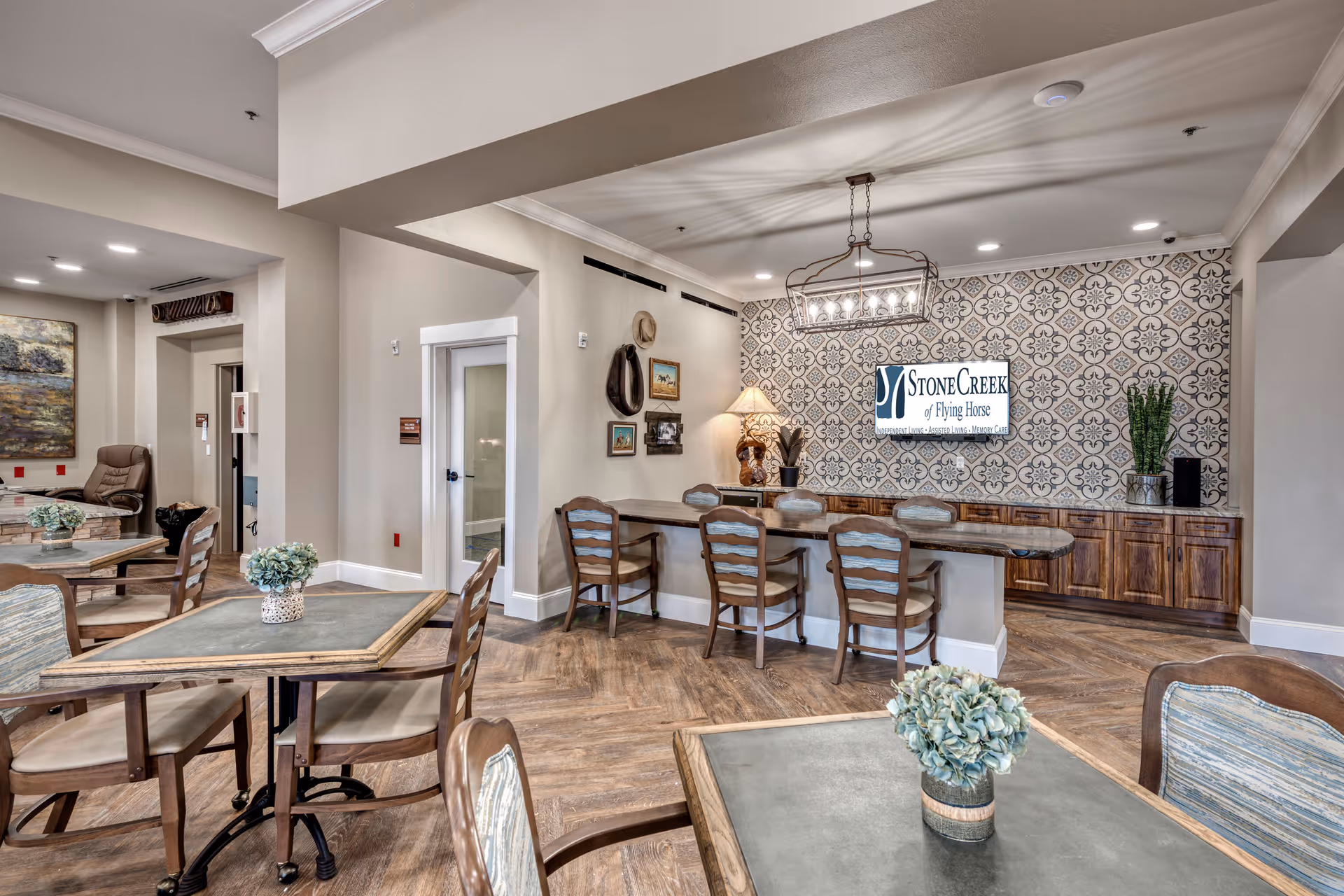 Interior view of a senior living facility common area with several tables and chairs arranged for seating. A counter with four chairs is positioned against a wall with decorative patterned wallpaper and a sign that reads 'StoneCreek of Flying Horse'. The room features wooden flooring, soft lighting, and decorative plants on the tables.