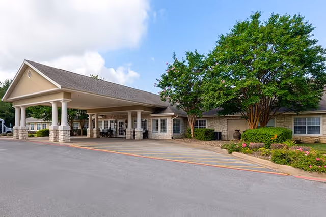 Front exterior view of Brookdale Tyler South facility showing a covered entrance with columns, stone facade, windows, and landscaped greenery including trees and bushes under a partly cloudy sky.