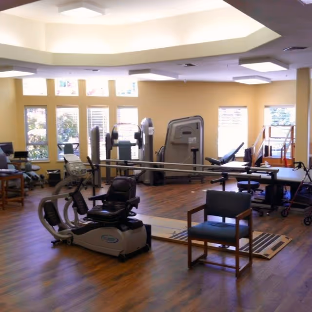 A spacious exercise room with various fitness equipment including a recumbent bike, weight machines, and parallel bars for physical therapy. The room has large windows allowing natural light to fill the space, wooden flooring, and a few chairs placed around the area.