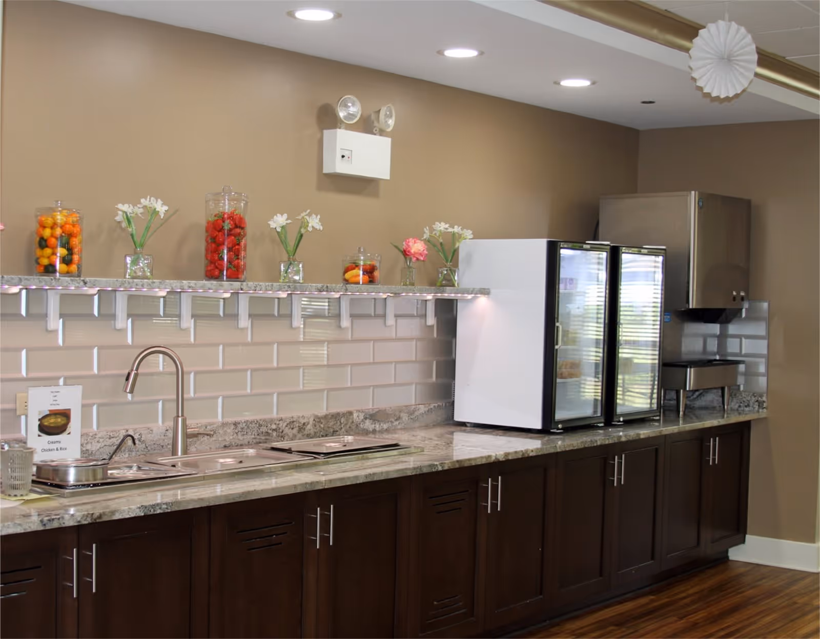 A kitchen area with a granite countertop, dark wooden cabinets, a stainless steel sink with a faucet, two glass-door refrigerators, and a stainless steel warming tray. Above the counter is a shelf holding jars of colorful fruits and small vases with white and pink flowers. The backsplash is made of white subway tiles, and the walls are painted beige.
