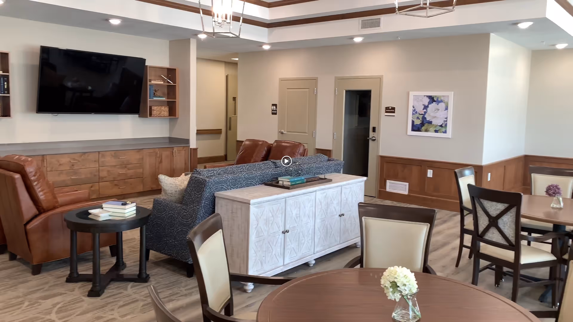 A cozy living room area in an assisted living facility featuring a mounted flat-screen TV above a wooden cabinet, two brown leather armchairs, a blue patterned sofa, and a white sideboard. There are round wooden tables with chairs around them, some with small flower vases. The room has light-colored walls, wood paneling, and a framed floral artwork on the wall.