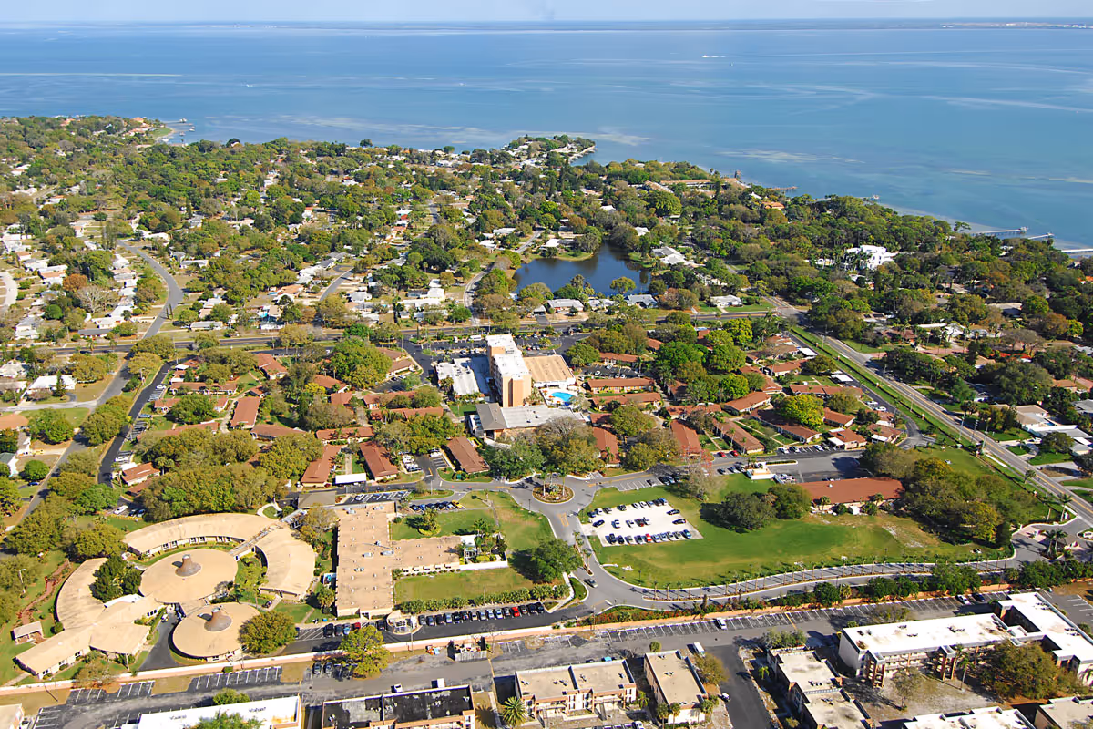 Aerial view of Westminster Suncoast senior living facility showing multiple buildings with red and beige roofs, surrounded by green trees and lawns, with a body of water in the background and a clear blue sky.