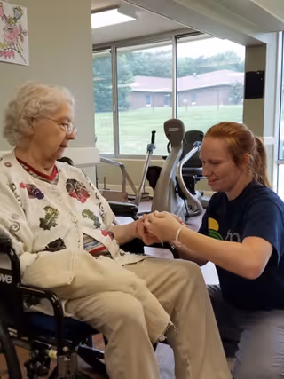An elderly woman sitting in a wheelchair inside a room with large windows. A younger woman is kneeling in front of her, holding and examining the elderly woman's hand. Exercise equipment is visible in the background.