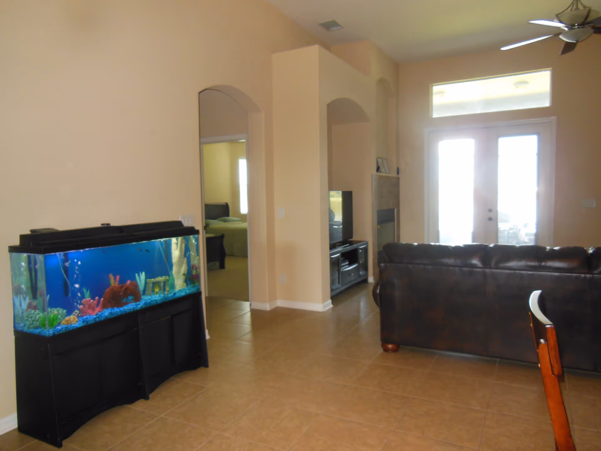 Interior view of a living room with a large fish tank on the left, a brown leather couch facing a TV and fireplace, and a doorway leading to a bedroom with a bed visible. There are double glass doors letting in natural light and a ceiling fan above.