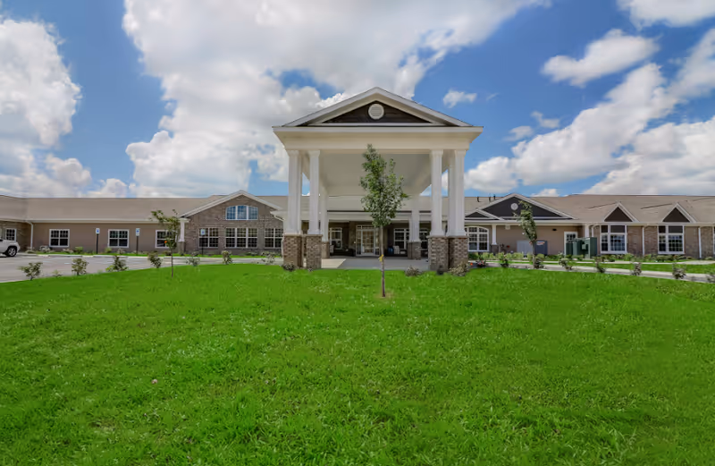 Front exterior view of Springvale Assisted Living & Memory Care building with a large covered entrance, green lawn, small trees, and a partly cloudy sky.