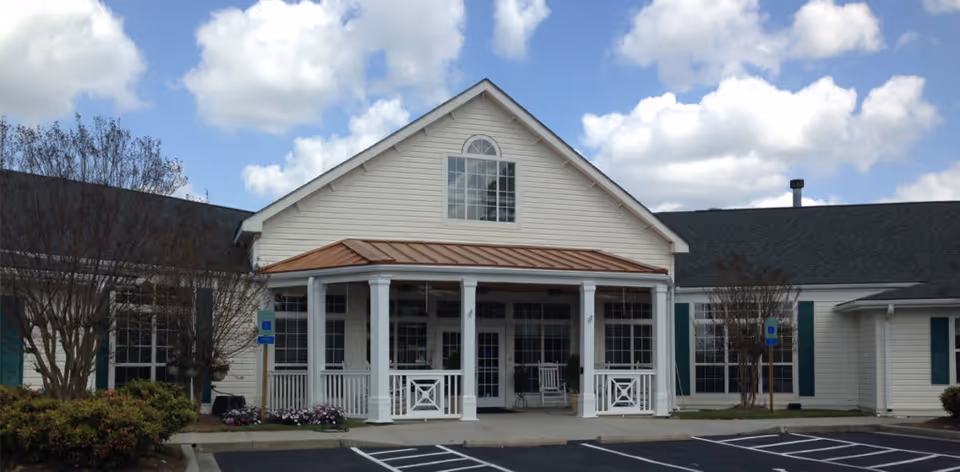 Front entrance of a light-colored senior living building with a covered porch, white columns, rocking chairs, and adjacent parking under a partly cloudy sky.
