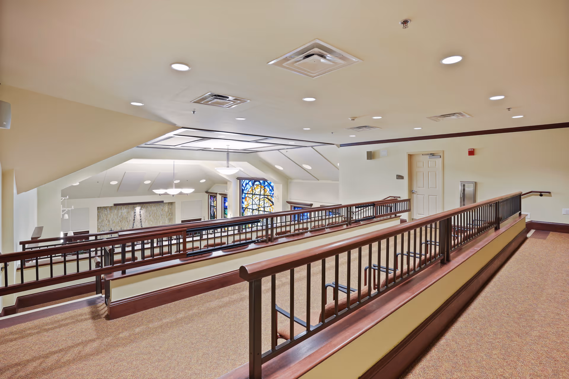 Interior view of a senior living facility showing a carpeted hallway with wooden handrails and metal balusters overlooking a lower level with stained glass windows and ceiling lights.