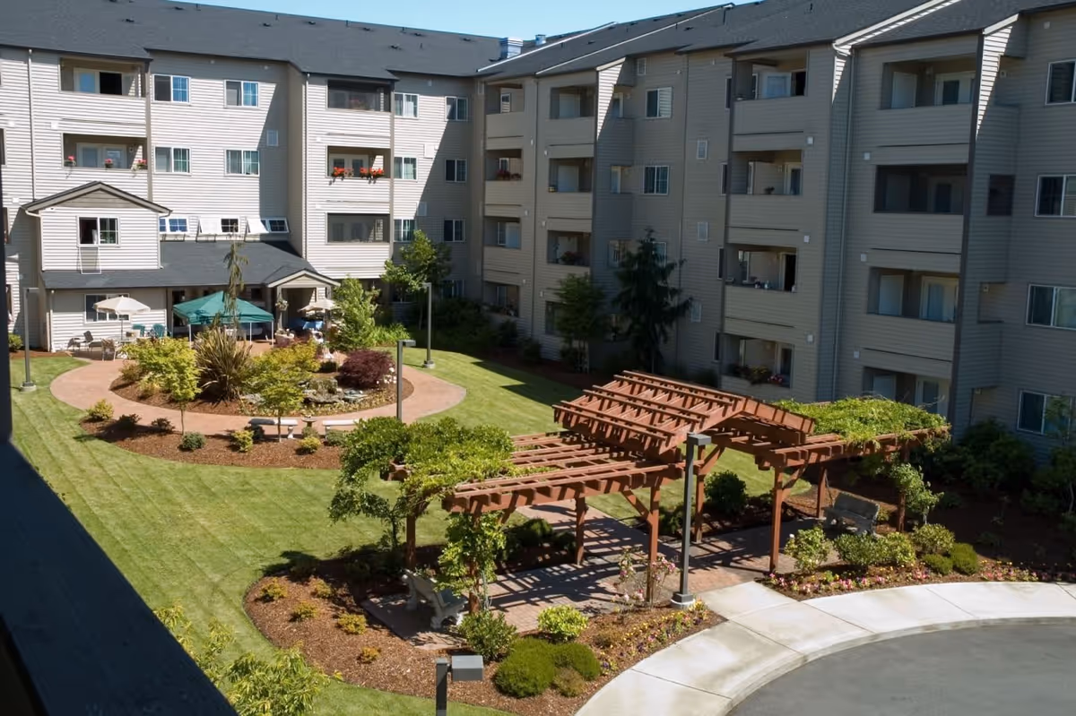 Courtyard garden with a wooden pergola, seating areas, umbrellas and landscaping surrounded by a multi-story senior living building.