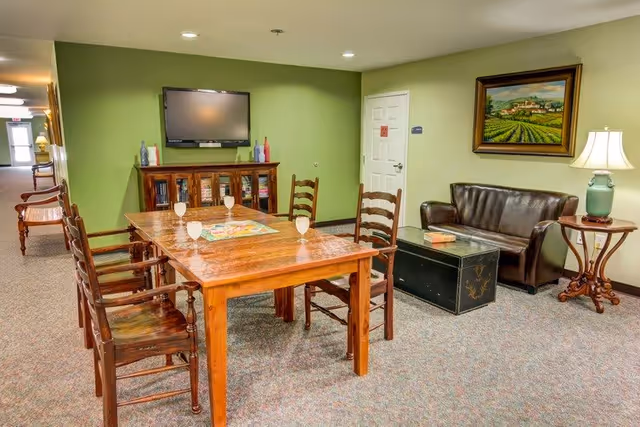 A cozy common area with a wooden table and four chairs, each with a glass of water on the table. There is a flat-screen TV mounted on a green wall above a wooden cabinet with glass doors. To the right, there is a brown leather couch, a black chest used as a coffee table, and a round wooden side table with a lamp. A framed painting of a vineyard hangs on the wall above the couch. The room has carpeted flooring and recessed ceiling lights.