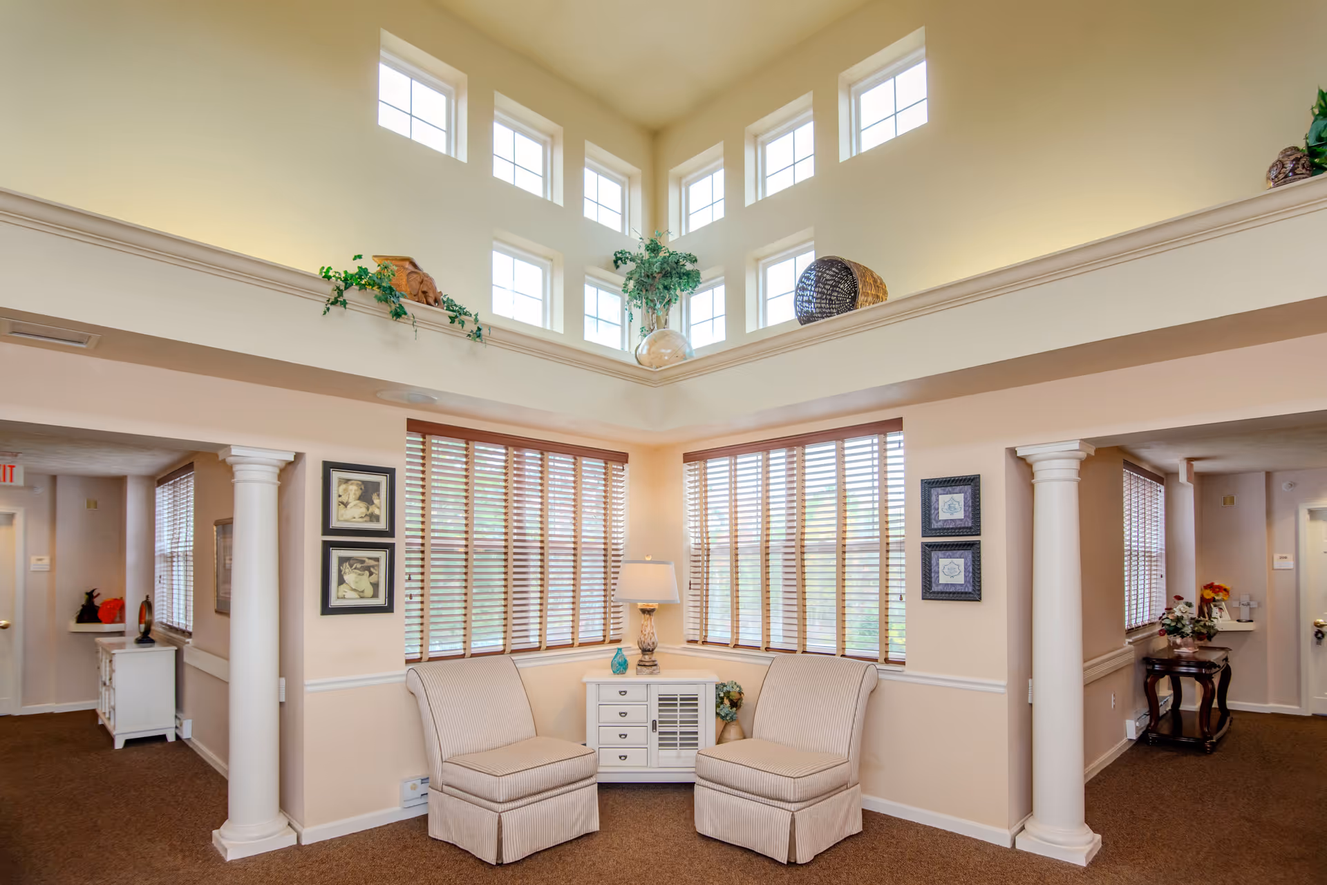 A bright and airy sitting area in a senior living facility with two beige upholstered chairs facing each other, a small white cabinet with drawers and a lamp between them, large windows with wooden blinds, and decorative plants and artwork on the walls. The space features high ceilings with multiple small square windows near the top and white columns framing the seating area.