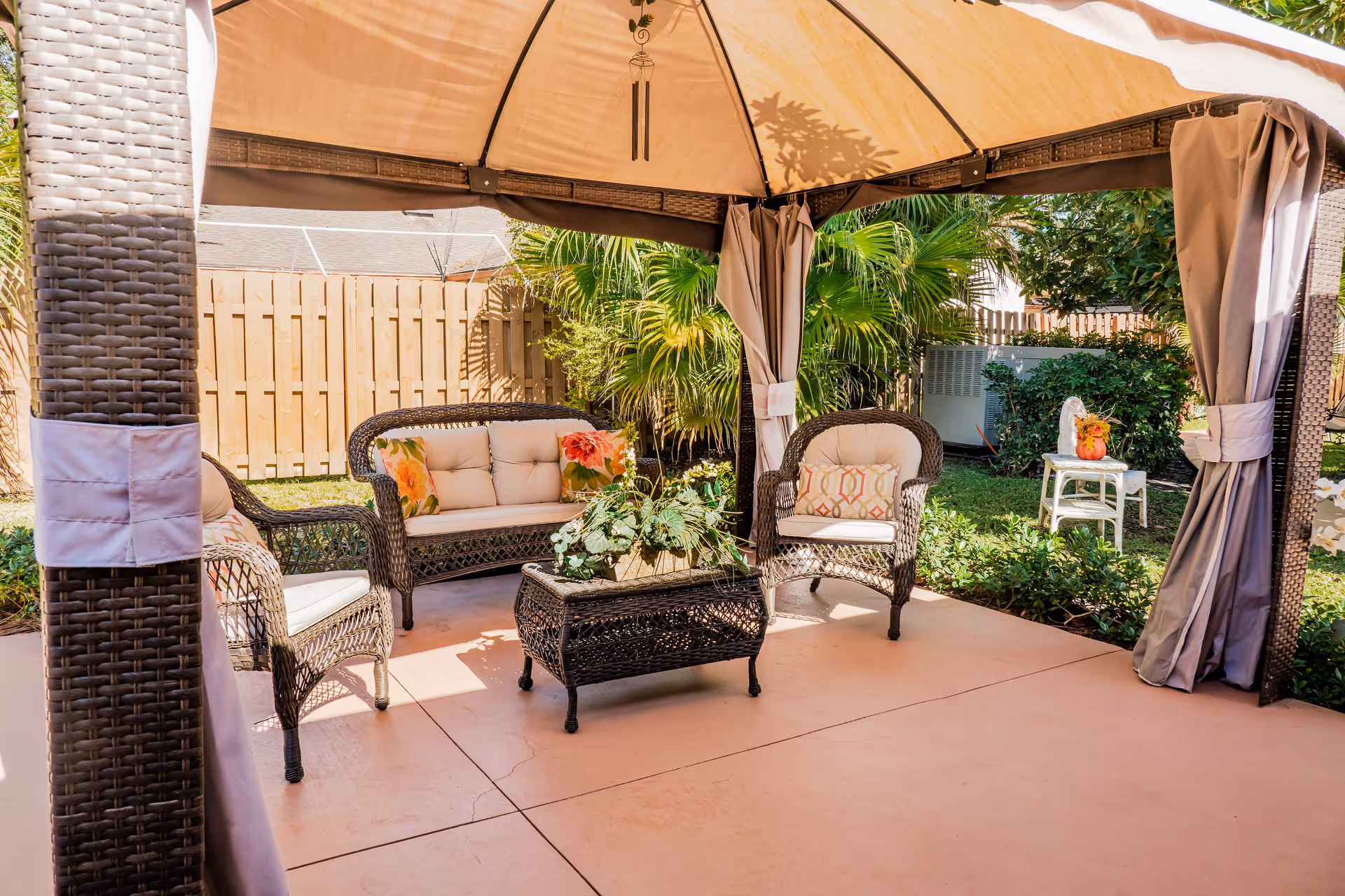 Outdoor seating area under a canopy with wicker furniture including a loveseat and two chairs with cushions and floral pillows, a wicker coffee table with a plant centerpiece, surrounded by greenery and a wooden fence in the background.