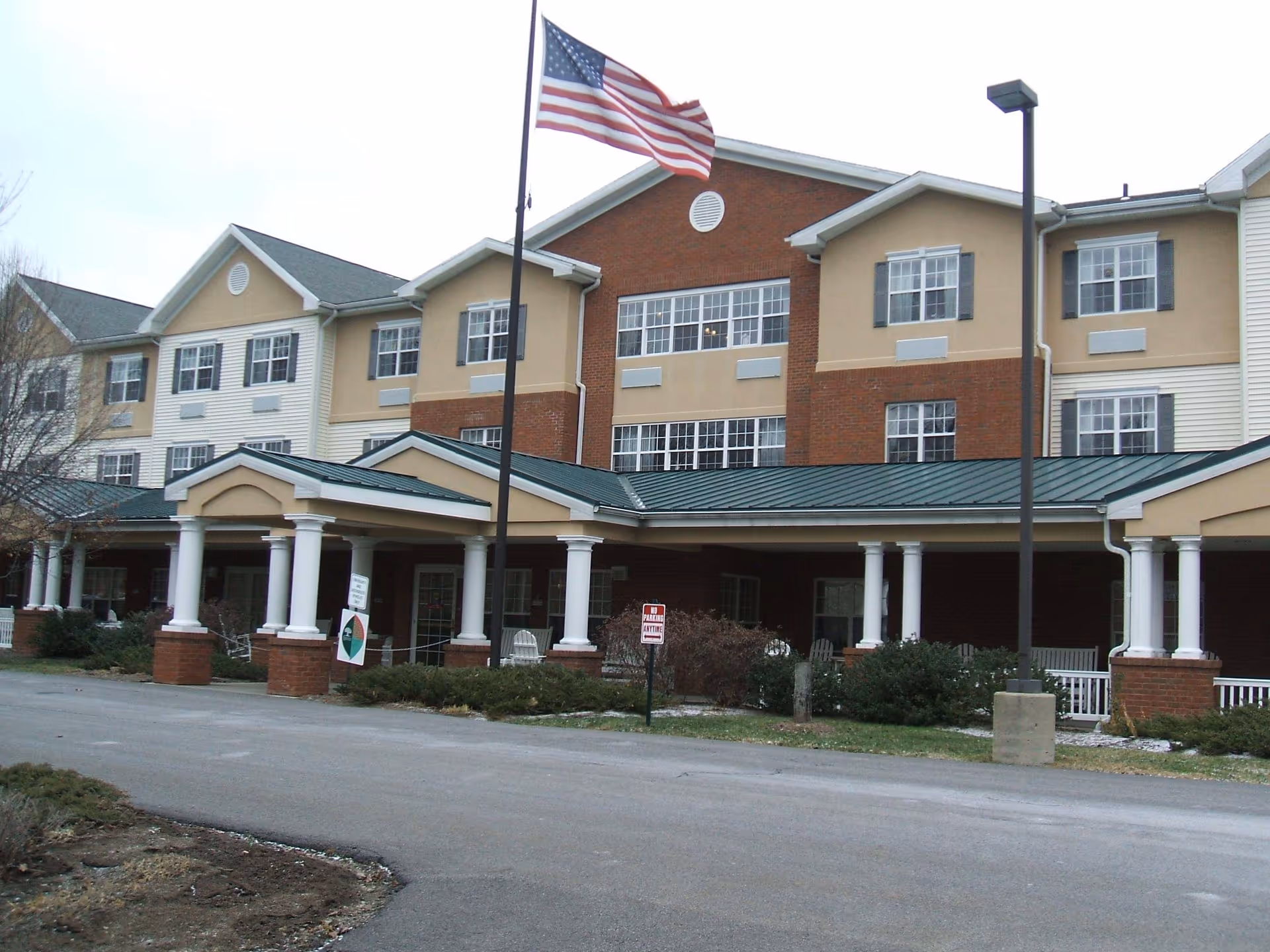 Exterior view of Clarks Summit Senior Living building with a covered entrance supported by white columns, an American flag on a flagpole, and a paved driveway in front.