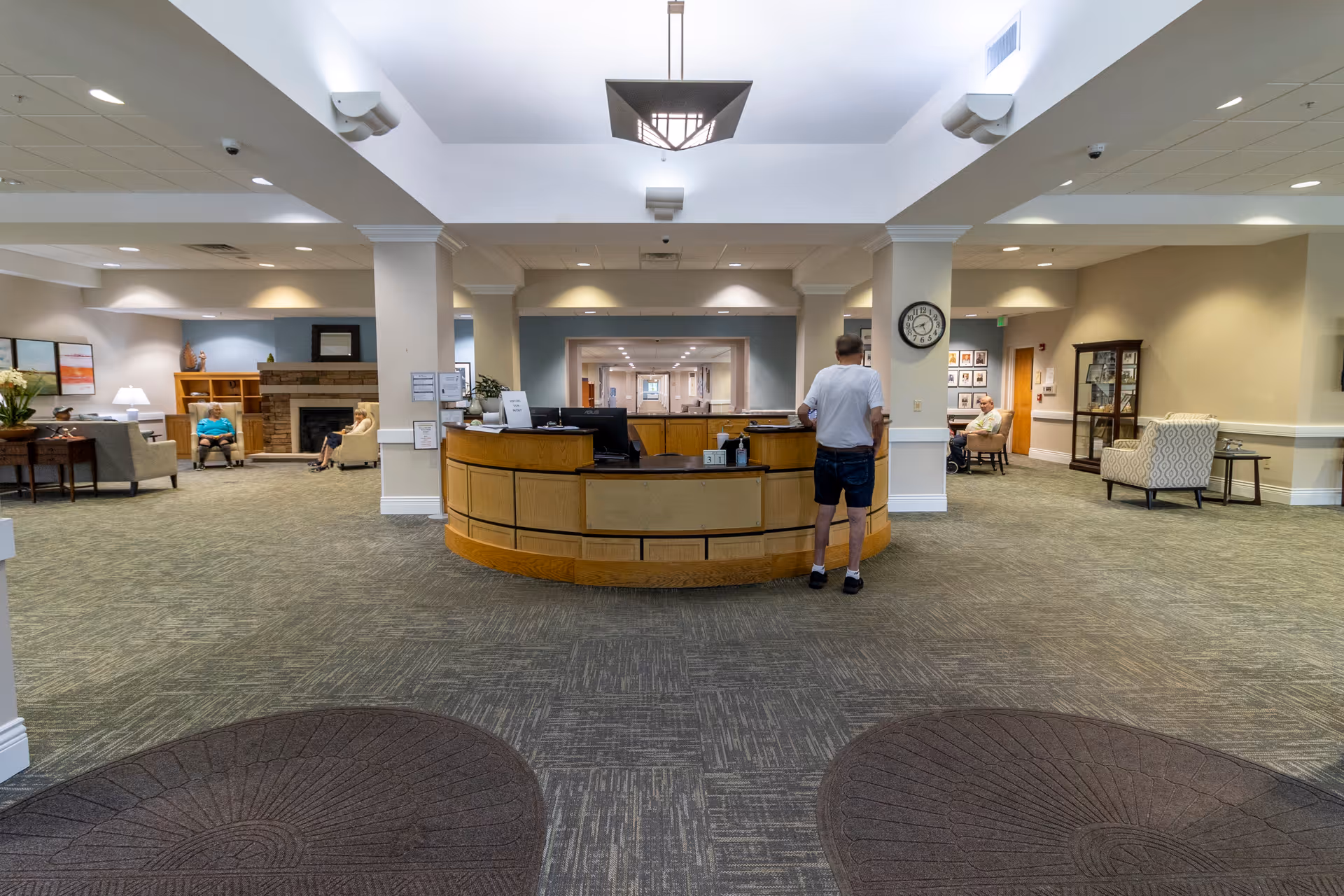 Interior view of a senior living facility lobby with a central reception desk. A man in casual clothes stands at the desk. The lobby has carpeted floors, comfortable seating areas with armchairs, a fireplace, and soft lighting. There are a few elderly people seated in the background, and a clock is mounted on a pillar near the reception desk.