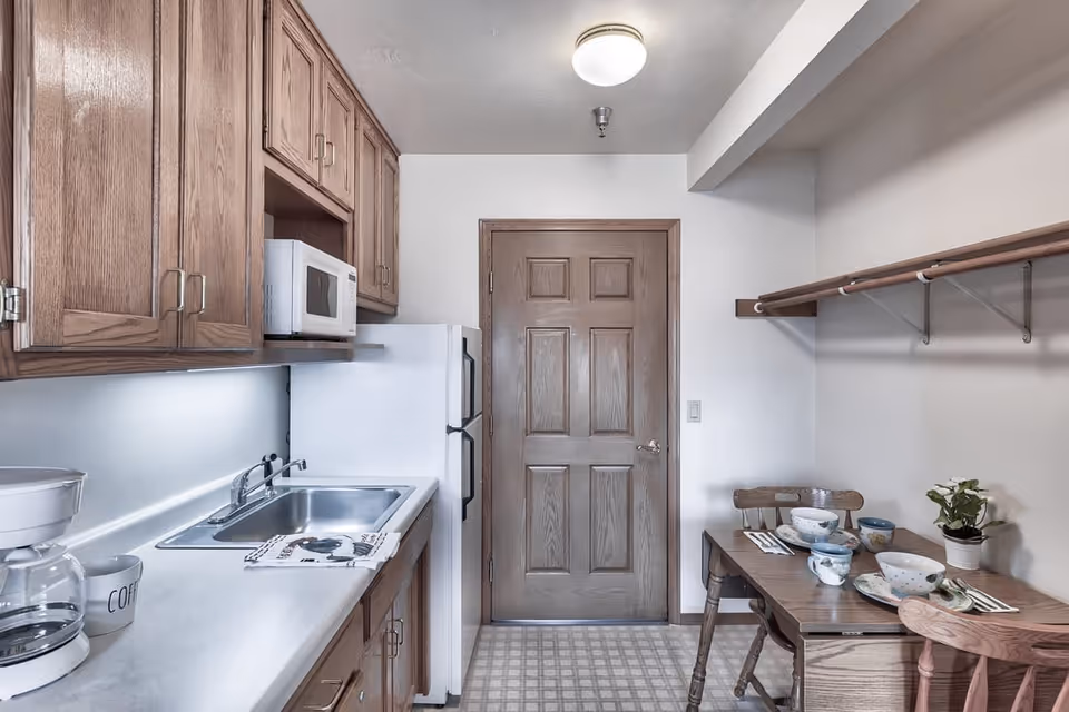 A small kitchen area with wooden cabinets, a white refrigerator, microwave, and coffee maker on the left side. On the right side, there is a wooden table set with bowls, cups, and utensils, accompanied by two wooden chairs. A wooden door is centered at the back of the room.