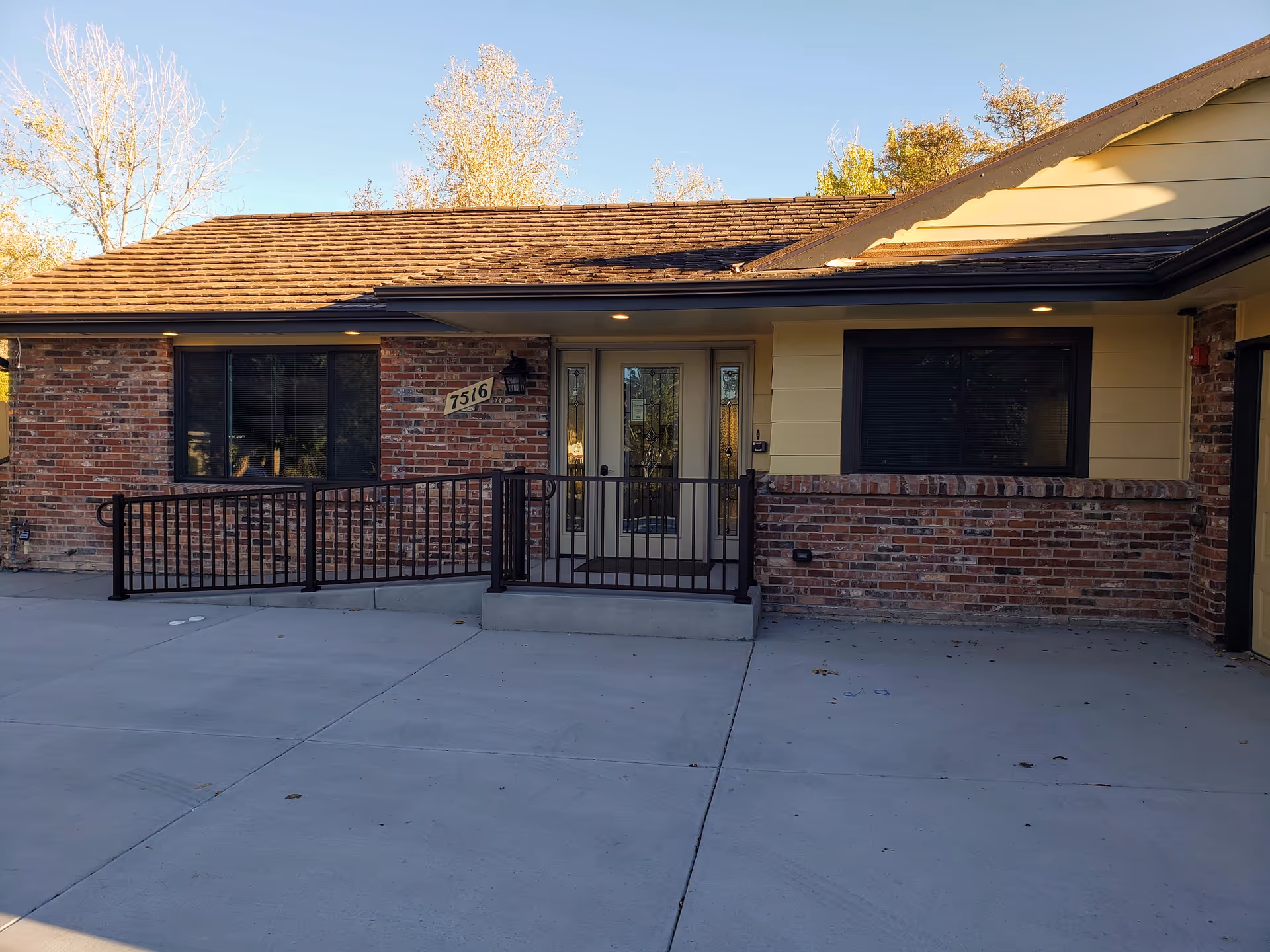Front exterior view of a single-story assisted living facility building with brick and yellow siding, a glass front door with sidelights, two windows, a black metal railing, and a concrete driveway. The building number 7516 is displayed near the door.
