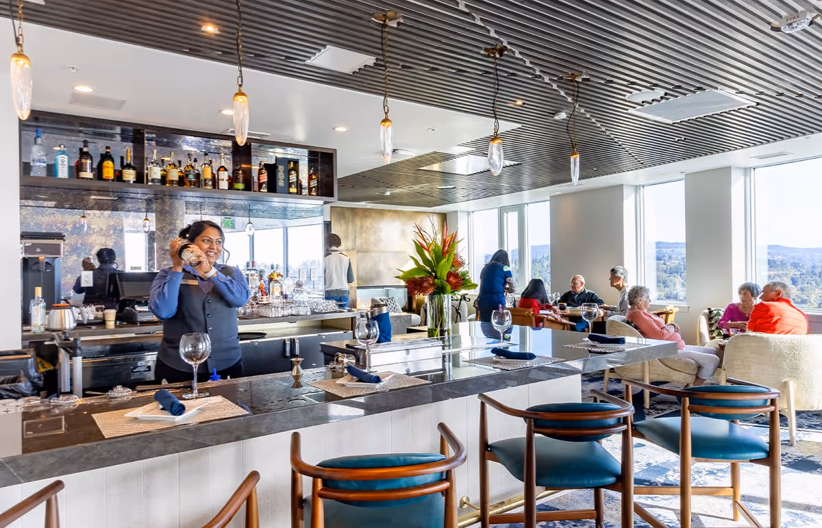 A bright and modern bar area in a senior living facility with a bartender shaking a cocktail shaker behind the counter. Several bottles of liquor are displayed on shelves behind the bar. In the background, elderly residents are seated and engaged in conversation near large windows with a scenic view outside. The space features stylish pendant lighting, comfortable seating, and a vase with flowers on the bar counter.