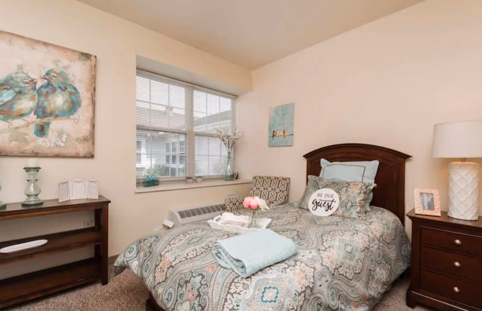 A cozy single-occupancy bedroom with a patterned quilt, a 'Be Our Guest' decorative pillow, wooden headboard, nightstand with lamp, and a window.
