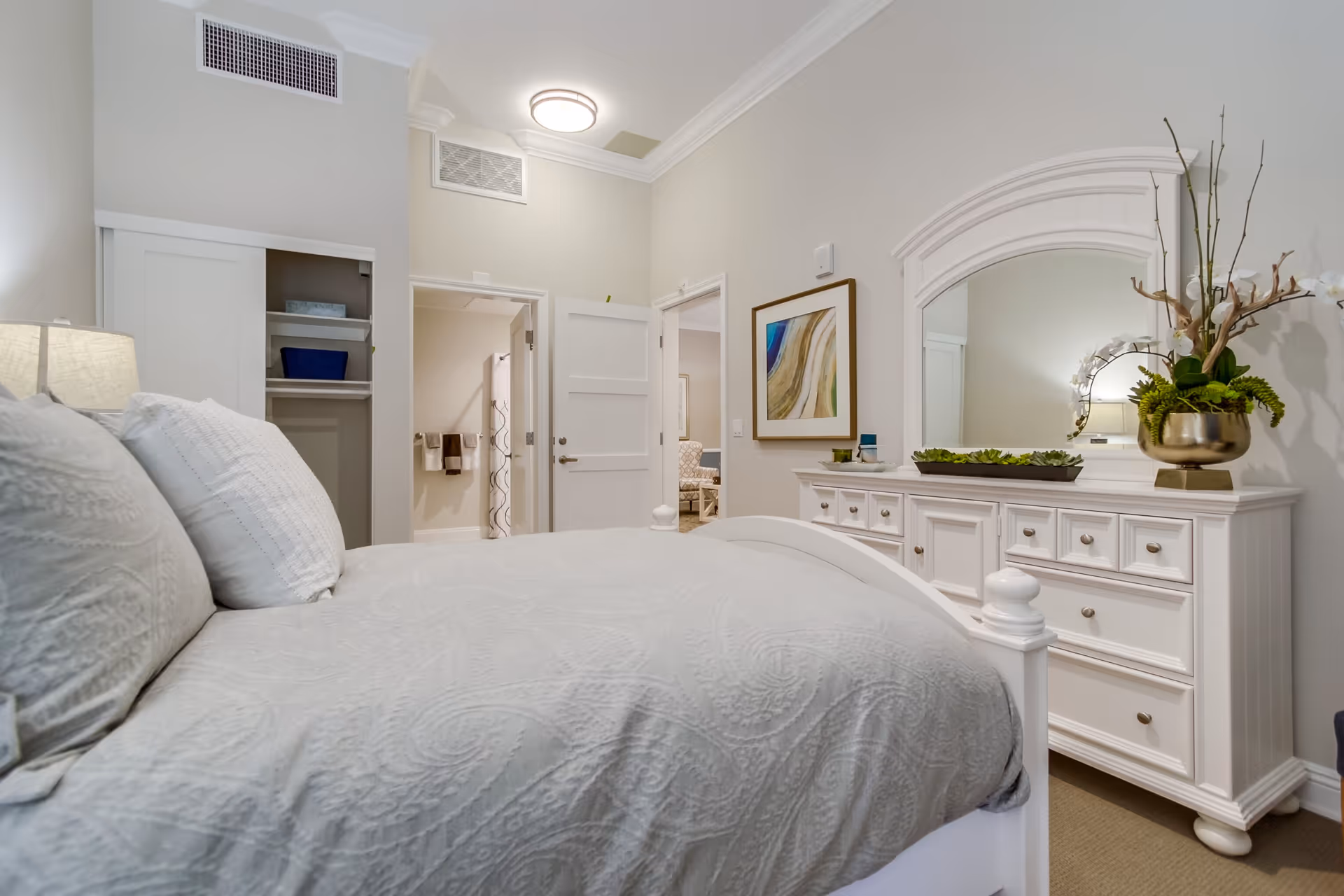 A neatly made bed with white bedding in a bedroom featuring a white dresser with a large mirror, a decorative plant, and a framed abstract painting on the wall. The room has light-colored walls, a closet with sliding doors, and an open door leading to another room.
