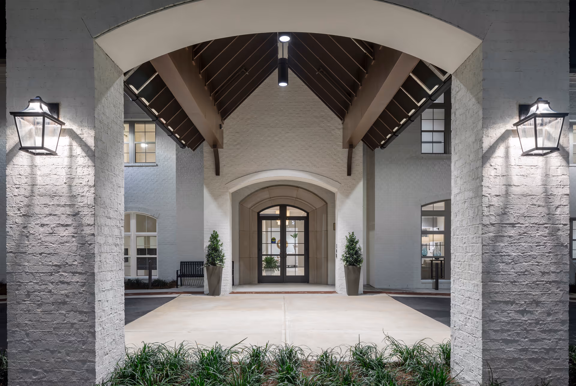 Entrance of The Blake at Chenal Valley facility at night, showing a covered driveway with two large pillars, wall-mounted lantern lights, potted plants on either side of the glass double doors, and a bench visible through the windows.