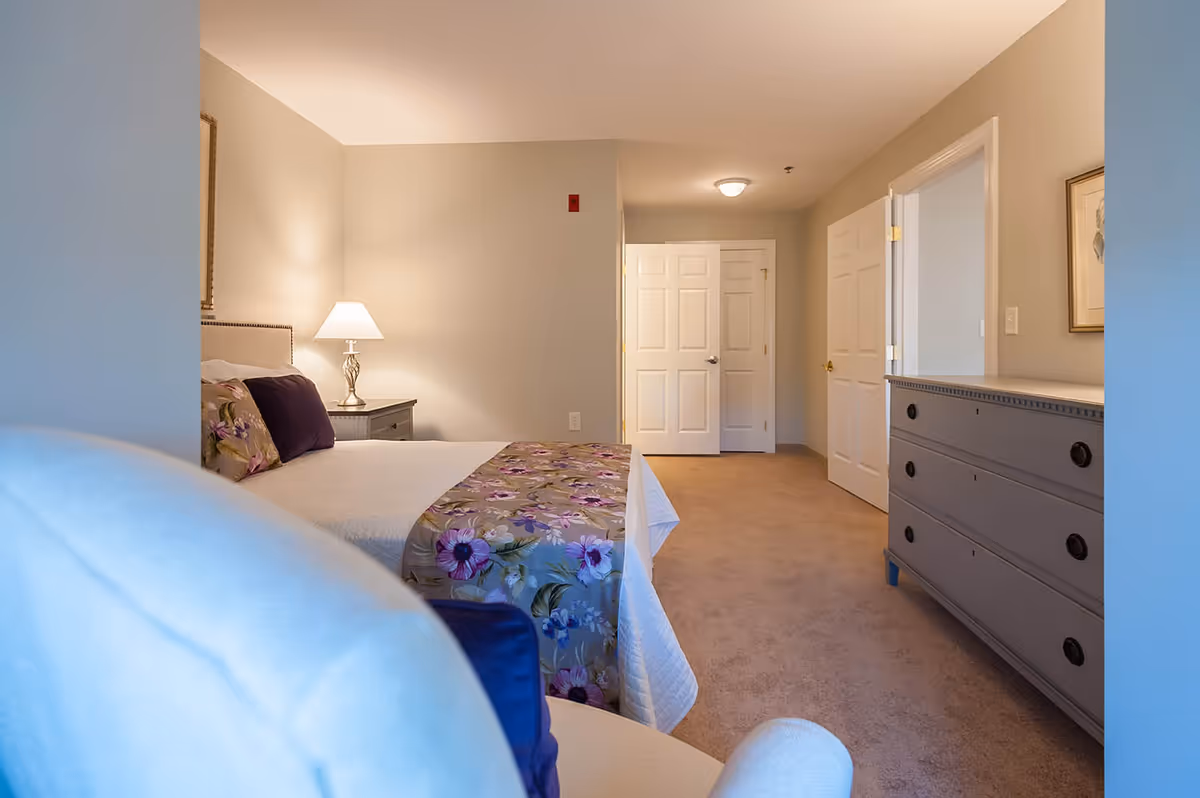 Well-lit furnished bedroom with a bed topped by a floral runner, a nightstand with lamp, a dresser, and open doors down a short hallway.