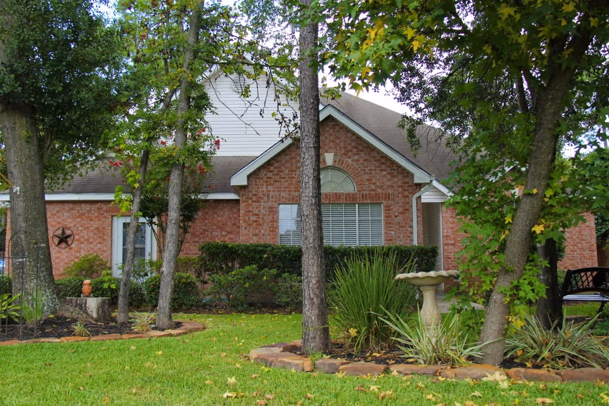 A brick cottage with white trim surrounded by trees and greenery. The front yard features a well-maintained lawn, shrubs, a birdbath, and a bench partially visible on the right side.