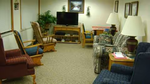 Carpeted senior living lounge with multiple chairs, couches, a TV, and side tables.