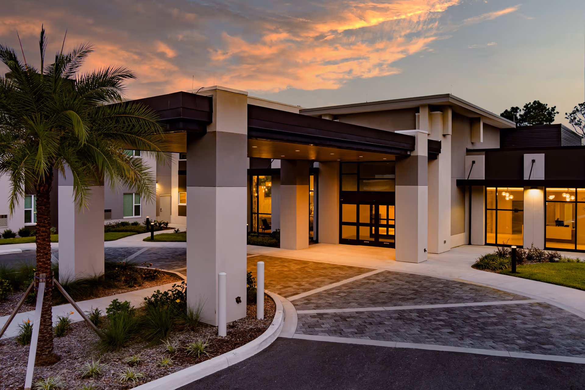 Exterior view of the Village on the Green facility entrance at sunset, featuring a covered drop-off area with large pillars, landscaped greenery including a palm tree, and warm interior lights visible through large windows.