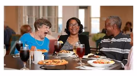 Three seniors sitting at a dining table in a senior living facility, smiling and enjoying a meal together with plates of food and glasses of beverages in front of them.