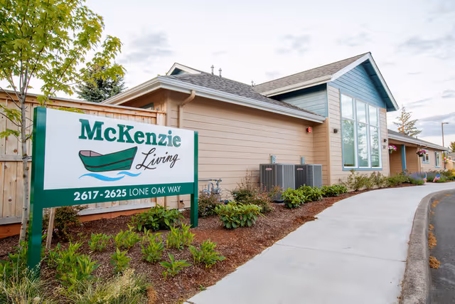 A McKenzie Living sign stands by landscaped beds in front of a single-story building with large windows and a curved sidewalk.