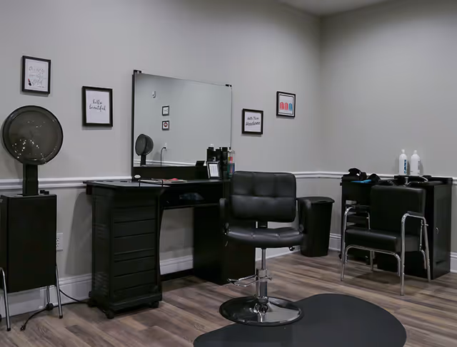 Small salon area with a styling chair, mirror, hooded dryer, and shampoo sinks against a light gray wall.