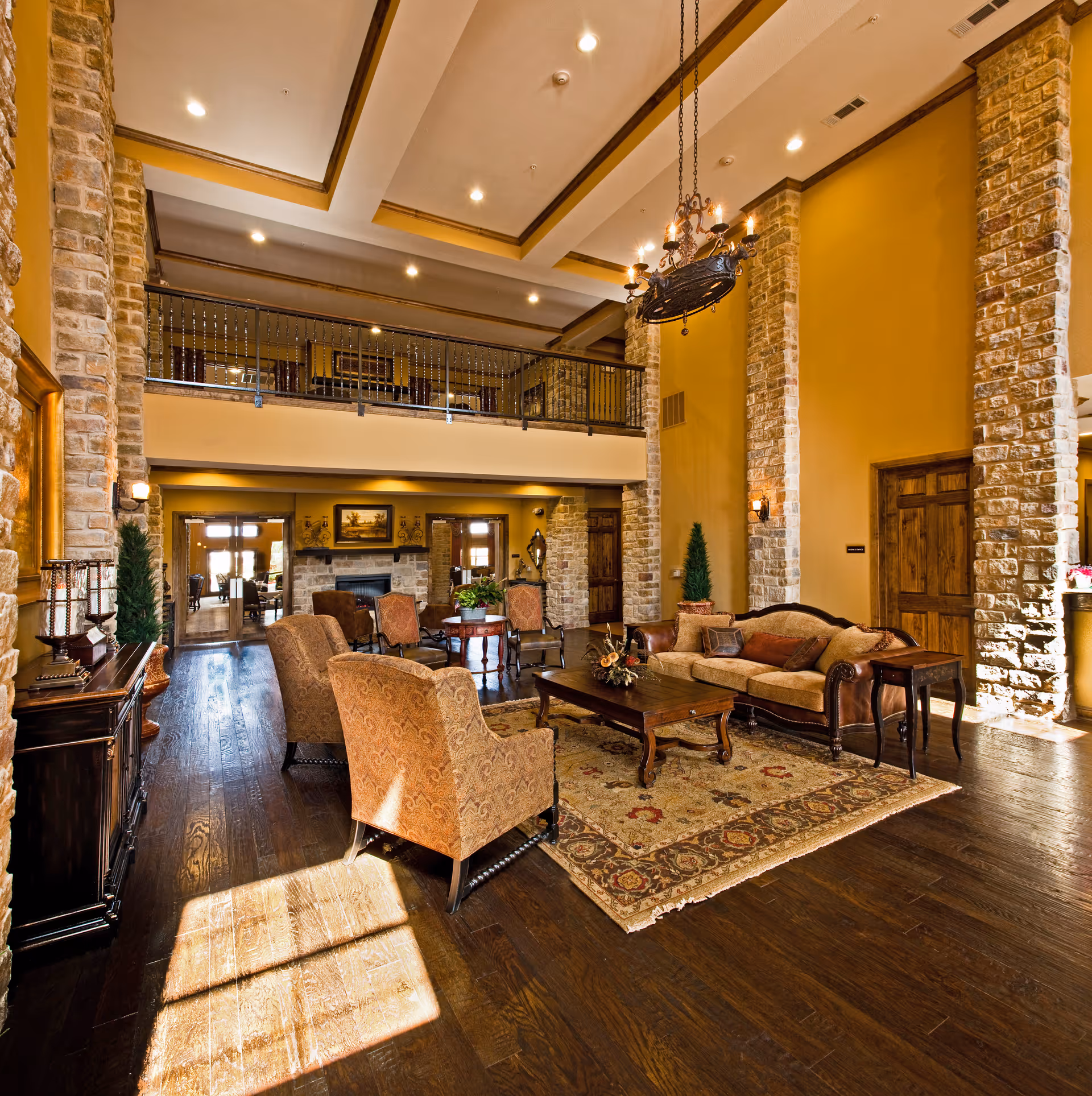 Spacious senior living facility common area with high ceilings, stone pillars, warm yellow walls, wooden floors, and elegant furniture including armchairs, a sofa, and a coffee table on a patterned rug. A chandelier hangs from the ceiling and there is a balcony railing on the upper level.