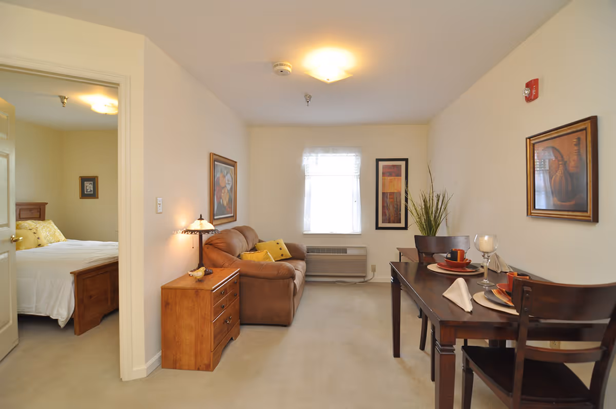 A cozy living and dining area in a senior living facility. The living area features a brown sofa with yellow cushions, a wooden side table with a lamp, and framed artwork on the walls. The dining area has a dark wooden table set for two with plates, cups, and napkins. A window with sheer curtains lets in natural light. To the left, an open door reveals a bedroom with a bed and framed pictures on the walls.
