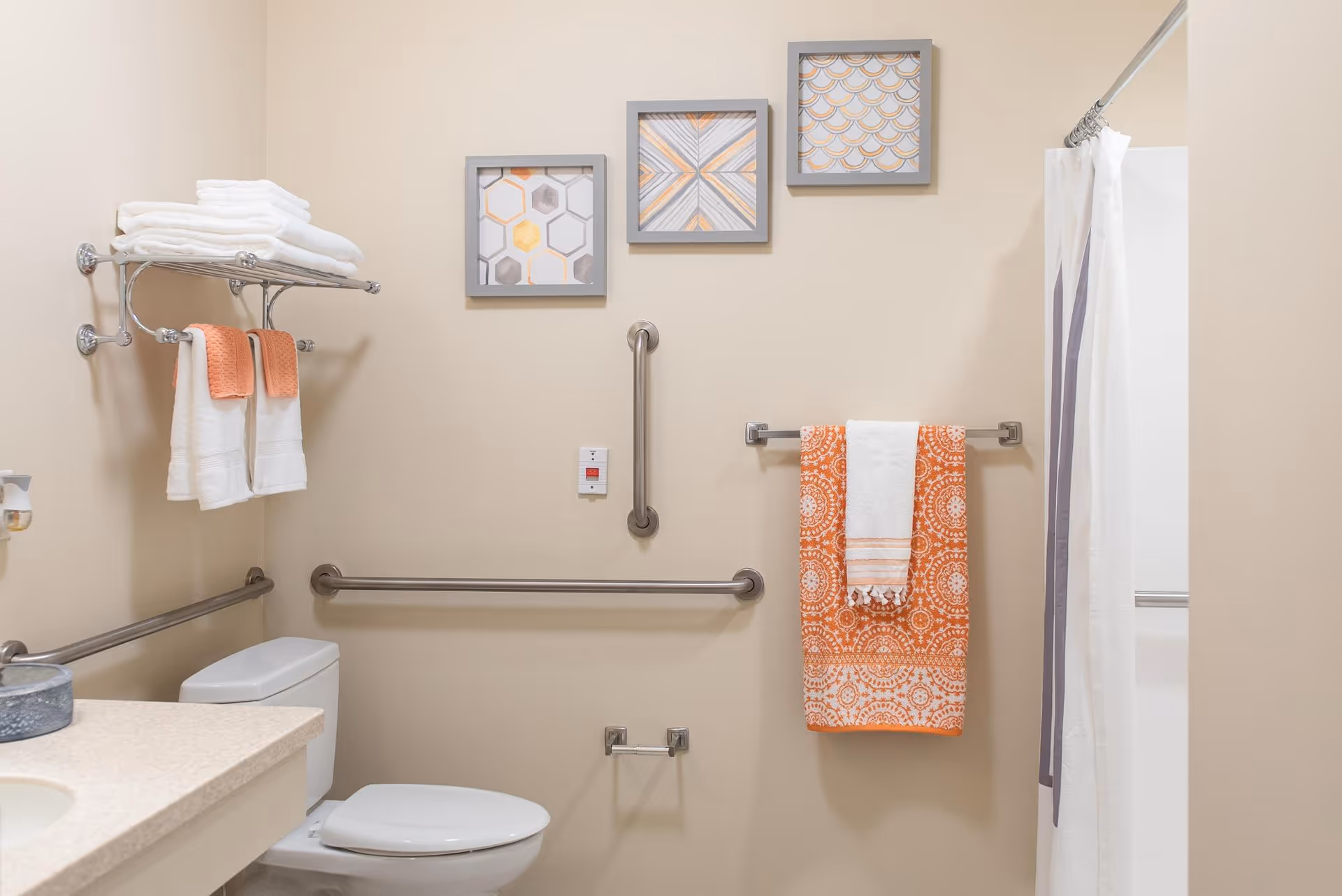 Accessible bathroom featuring a toilet, grab bars, towel racks with white and orange towels, a shower curtain, and framed wall art.