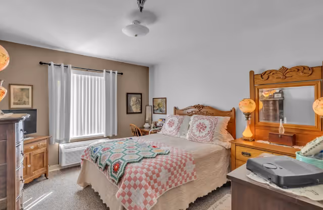 Sunlit bedroom featuring a decorative wooden bed with patchwork quilts, matching vintage dressers and a window with vertical blinds.