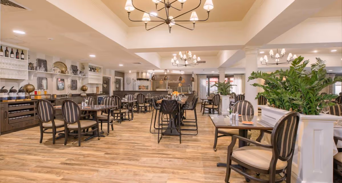 Spacious dining room with multiple tables and striped upholstered chairs, chandeliers, plants, and a serving counter.