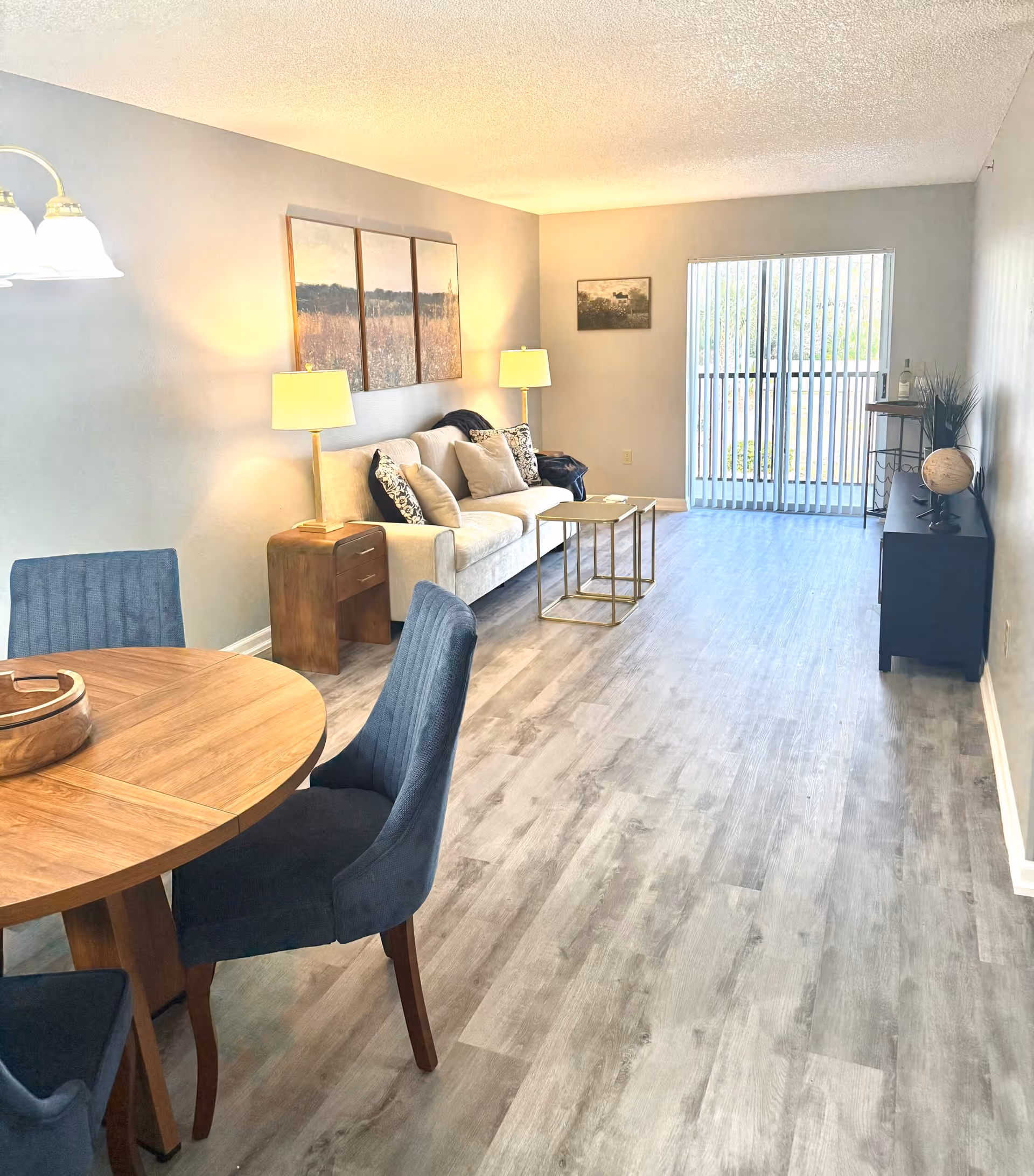 A bright living room and dining area with light gray wood flooring. The living room features a beige sofa with multiple cushions, two side tables with lamps, a small coffee table, and a black TV stand with a globe and decorative items. The dining area has a round wooden table with four dark upholstered chairs. Large vertical blinds cover a sliding glass door leading to a balcony or outdoor space. The walls are light gray with framed artwork hanging above the sofa and on the adjacent wall.